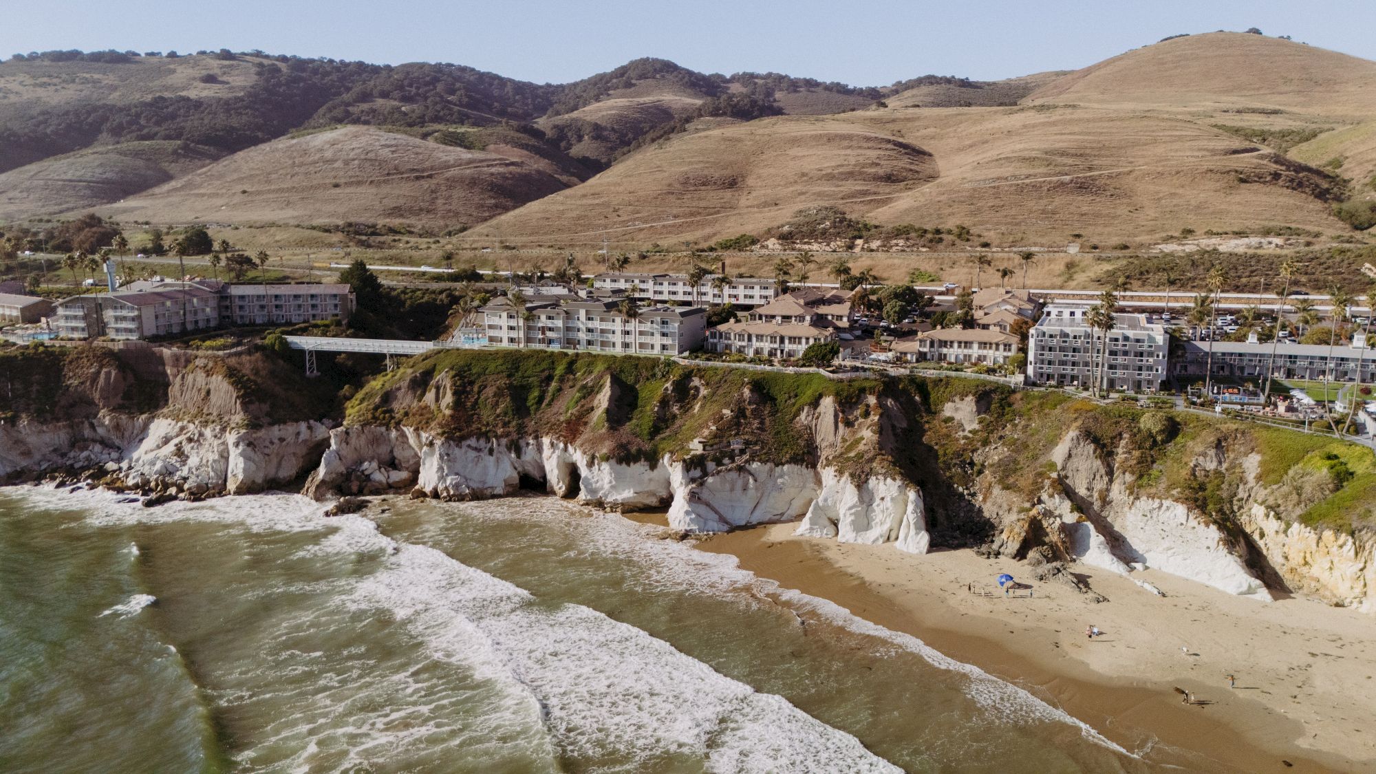 A coastal resort with apartment buildings by a sandy beach, waves crashing under rugged cliffs and rolling green hills.