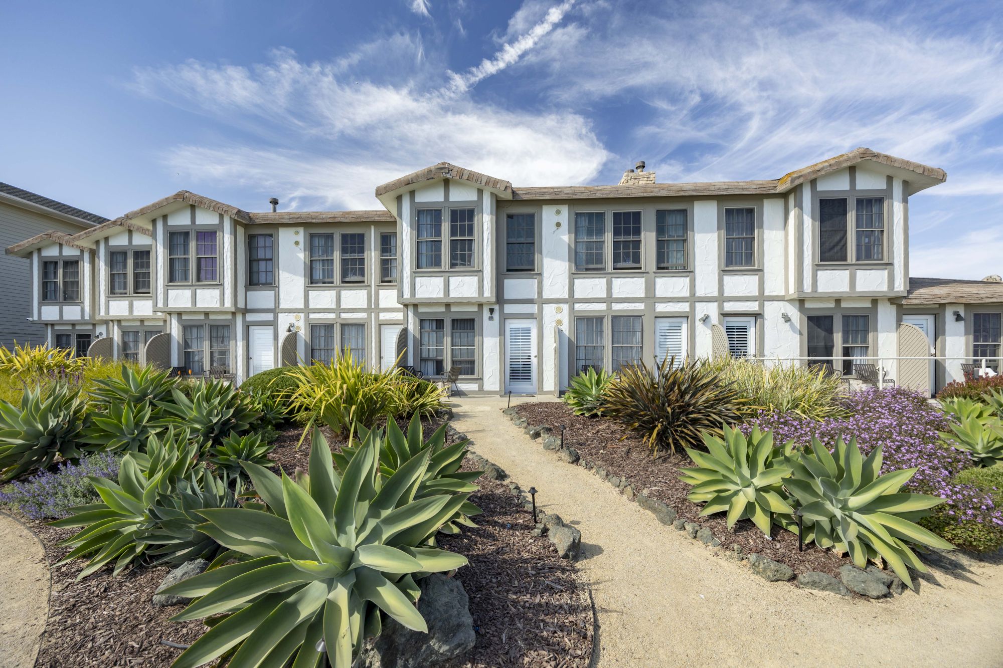 A bright two-story townhouse with dormers and bay windows, surrounded by succulent gardens and a sunny walkway under blue sky.