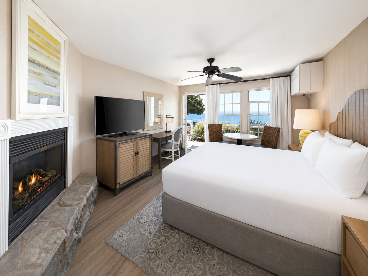 Modern hotel bedroom with a king bed, fireplace, TV, ceiling fan, and ocean-view windows and curtains.