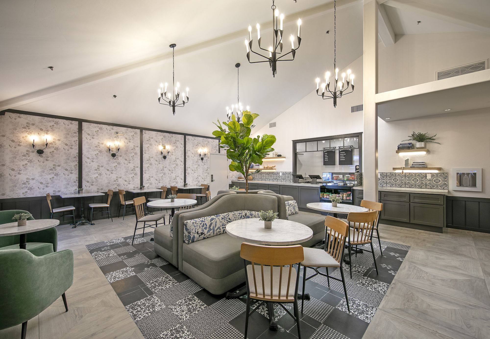 A modern cafe lounge with green sofas, round tables, hanging chandeliers, and a service bar in the back.