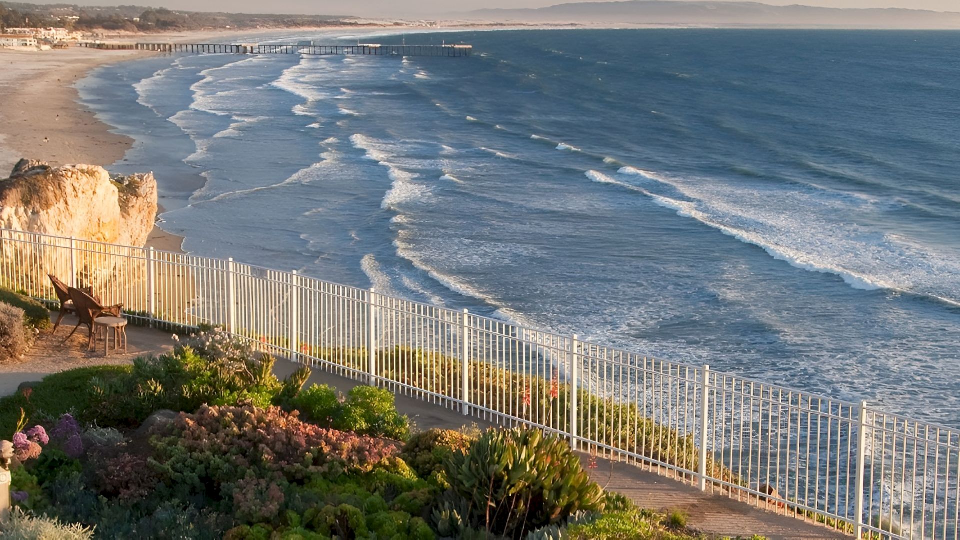 Ocean waves crash beside a wooden fence on a sunny coastal cliff, with a quiet sandy beach and homes in the distance.