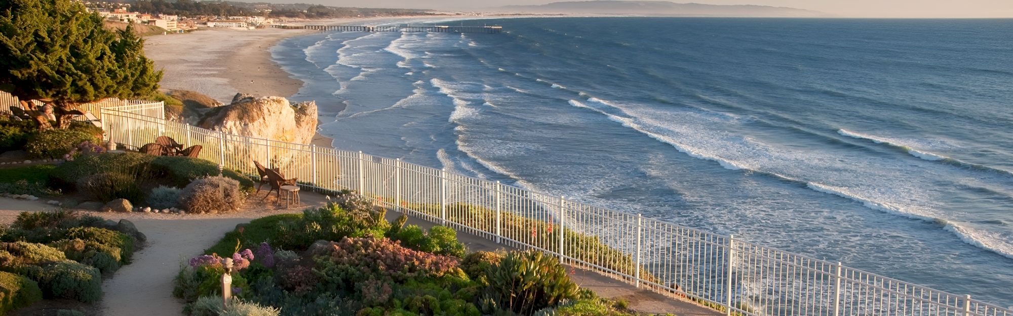 Ocean waves crash beside a wooden fence on a sunny coastal cliff, with a quiet sandy beach and homes in the distance.