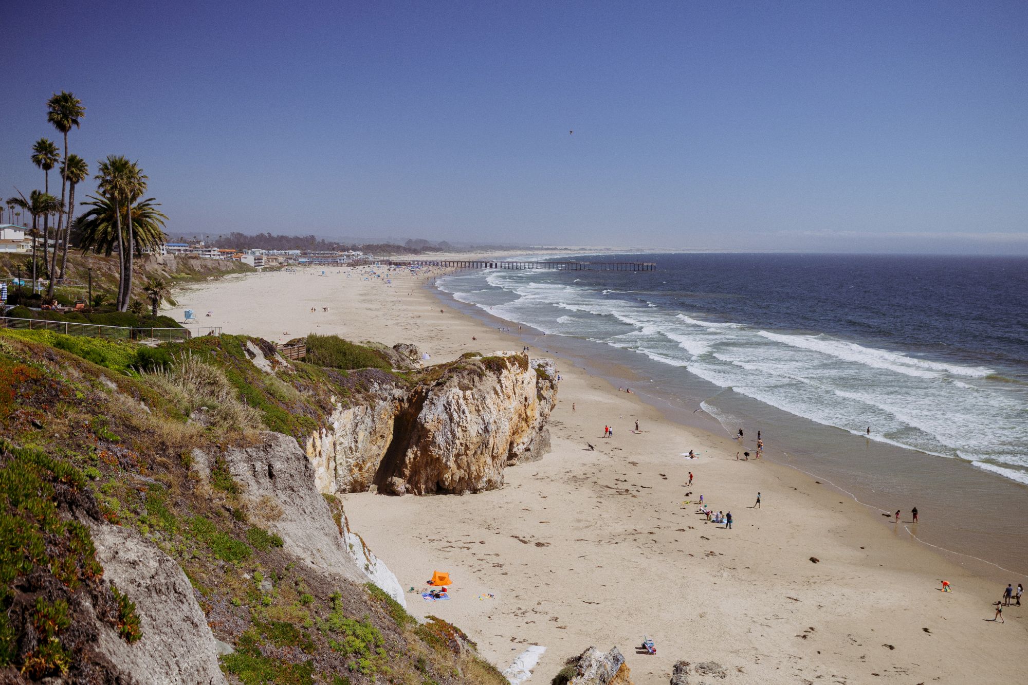 A sunny coastline with a sandy beach, crashing waves, palm trees, and people strolling near sea cliffs.