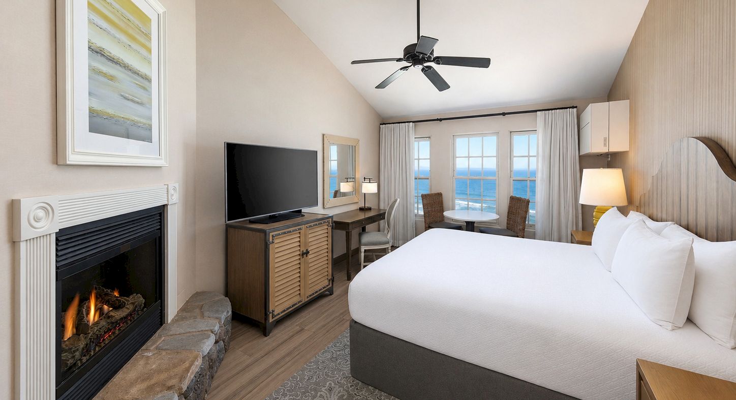 A bright hotel bedroom with a bed, TV, fireplace, ceiling fan, and large ocean-view windows.