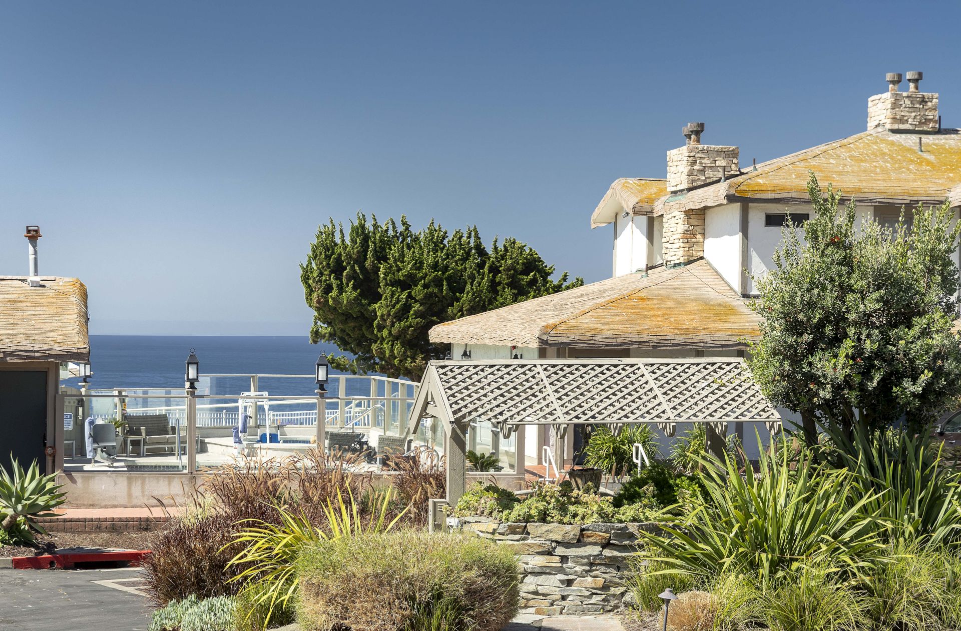 A seaside patio with lounge chairs, a white stone-roof building, trees and shrubs, and the ocean stretching to the horizon.