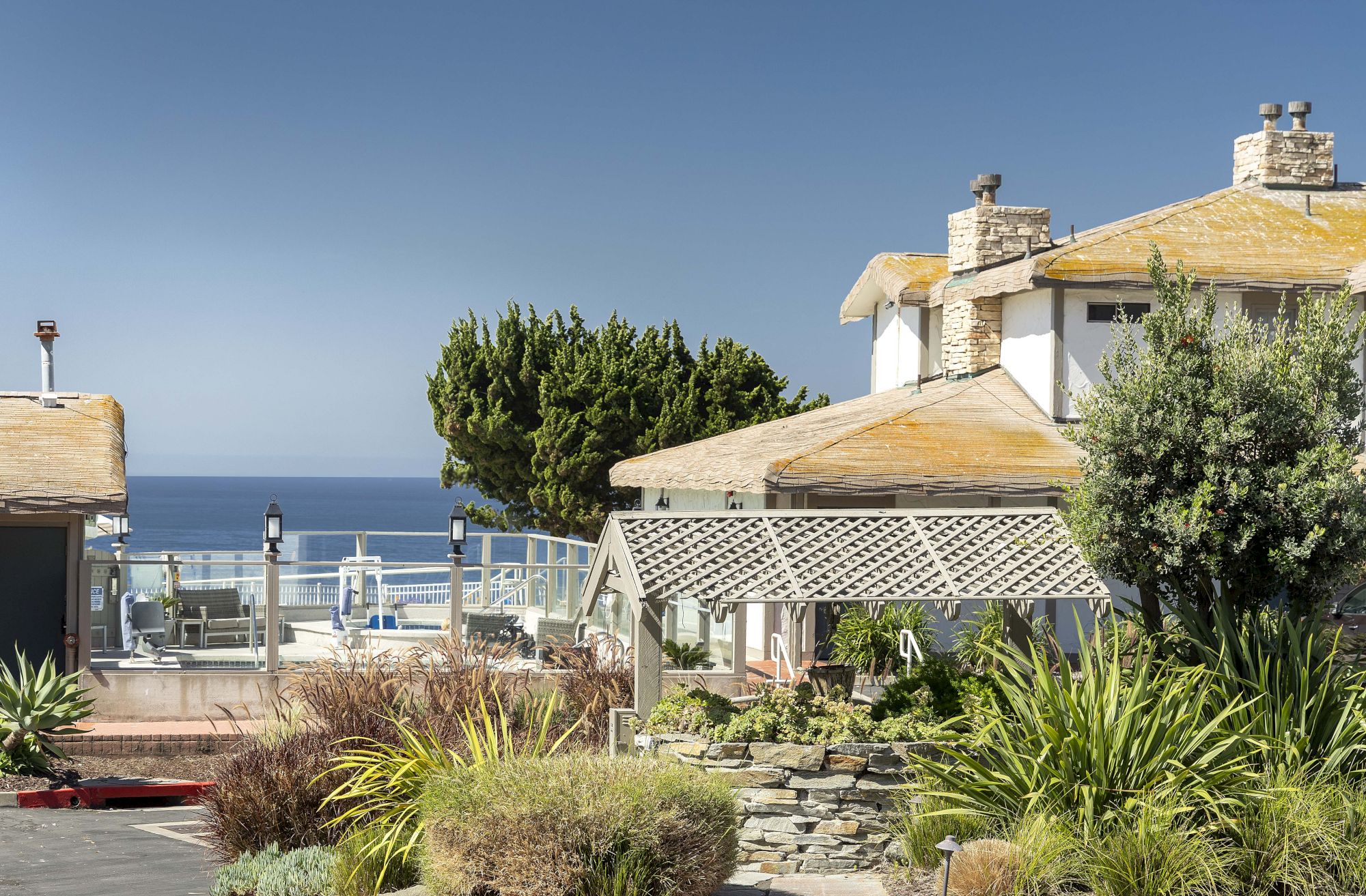 A seaside patio with lounge chairs, a white stone-roof building, trees and shrubs, and the ocean stretching to the horizon.
