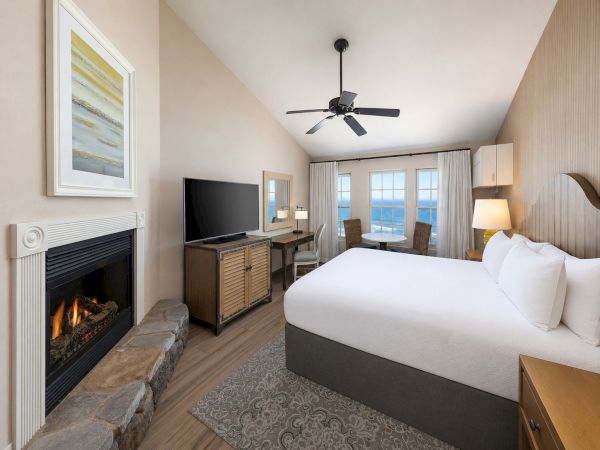 A modern bedroom with a bed, TV stand, fireplace, and ceiling fan, plus windows with curtains.