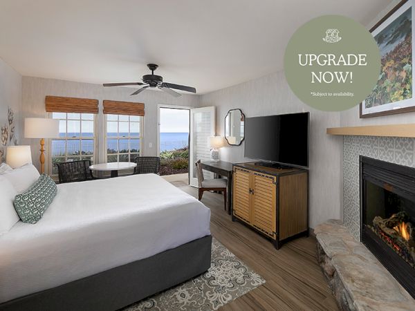 A modern hotel room with a large bed, TV, desk, fireplace, ocean-view window, and an &ldquo;UPGRADE NOW!&rdquo; banner.