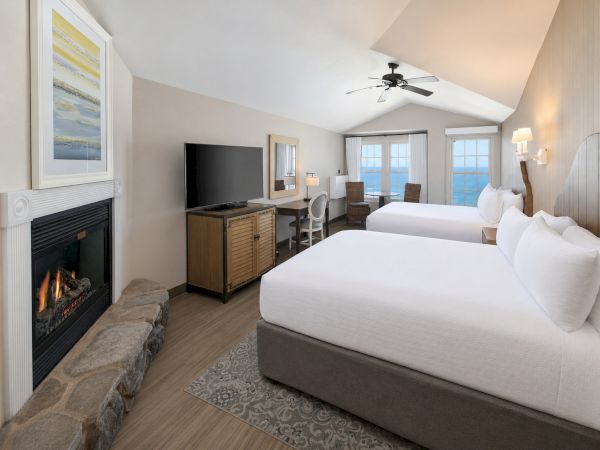 A bright hotel suite with two white beds, TV, fireplace, large windows, and a ceiling fan, creating a modern seaside feel.