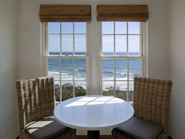 A sunny room with two wicker chairs and a round table, looking out through large windows at the ocean waves and sky.