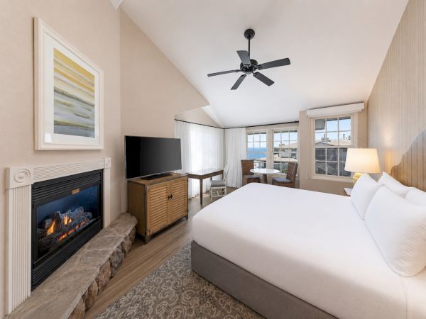 A modern hotel bedroom with a fireplace, TV, ceiling fan, large bed, and ocean-view windows.