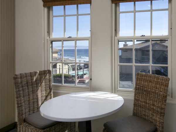 Two wicker chairs and a round table by large windows show ocean waves and coastal buildings outside.