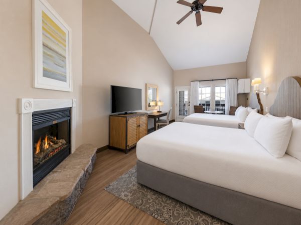 A bright hotel bedroom with two beds, a fireplace, TV, ceiling fan, and large windows.