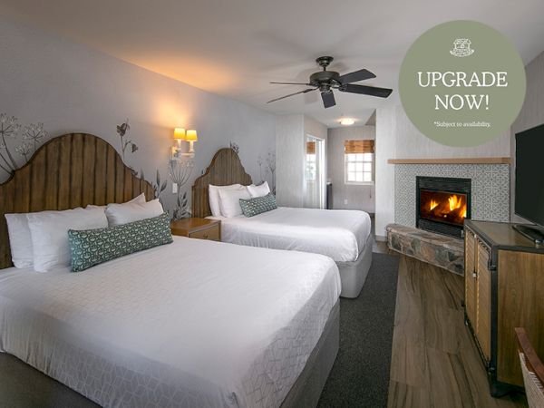 A hotel room with two beds, a fireplace, TV, and ceiling fan, plus an &ldquo;UPGRADE NOW!&rdquo; promo badge.