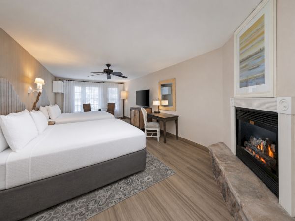 A bright hotel-style bedroom with two beds, a desk, TV, ceiling fan, and a lit stone fireplace.