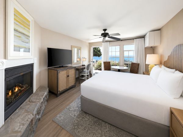 A modern hotel bedroom with a king bed, TV and fireplace, ceiling fan, and windows overlooking the ocean.
