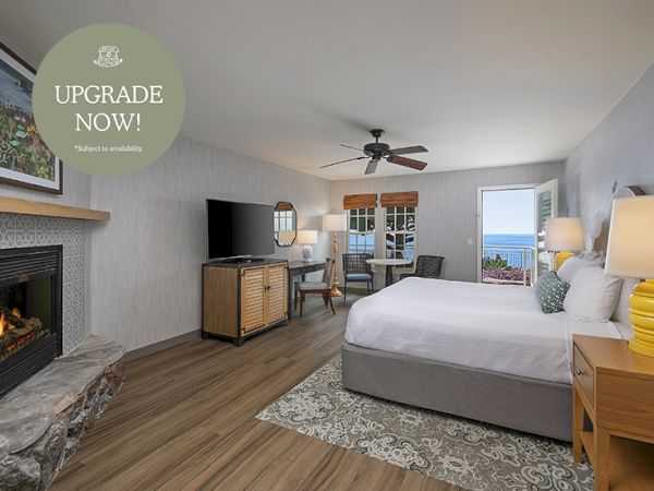 A bright hotel bedroom with a large bed, TV cabinet, ceiling fan, windows, and a stone fireplace.