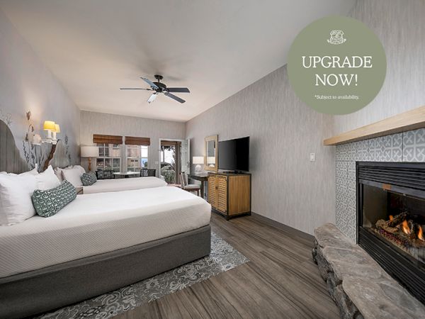 A cozy hotel room with a large bed, TV, fireplace, ceiling fan, and an &ldquo;UPGRADE NOW!&rdquo; promo sign.
