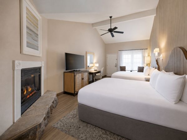 A bright hotel-style bedroom with a fireplace, ceiling fan, TV stand, lamps, and neatly made beds with pillows.