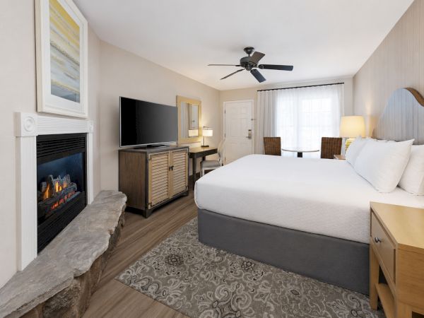 A hotel-style bedroom with a king bed, fireplace, TV on a cabinet, ceiling fan, and sheer curtains.