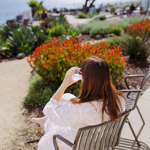 A woman in a pink dress sits on a patio chair, gazing at a vibrant flower garden and a tree in the distance.