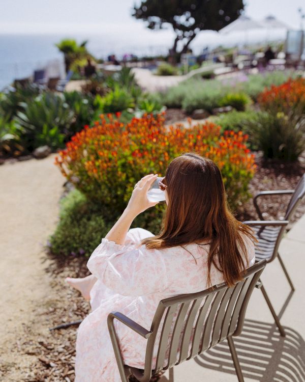 A woman in a pink dress sits on a patio chair, gazing at a vibrant flower garden and a tree in the distance.
