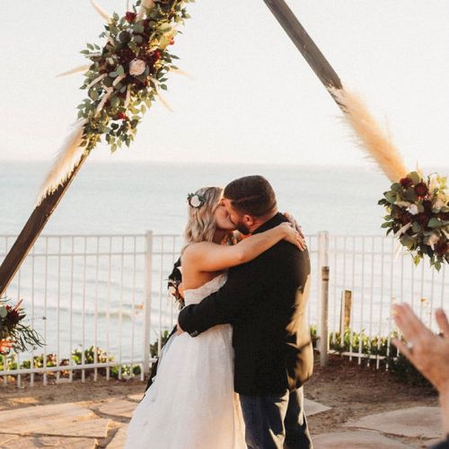 A couple kisses under a floral arch at a beach wedding ceremony, with candles, bouquets, and a decorative rug.
