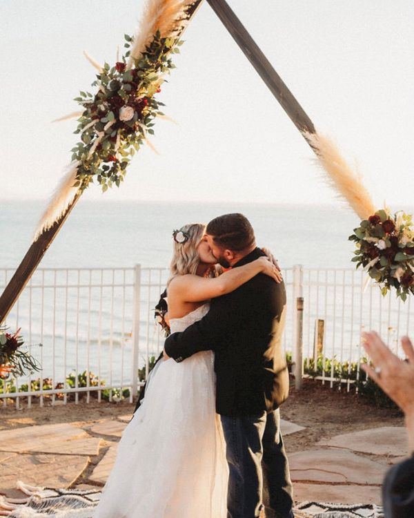 A couple kisses under a floral arch at a beach wedding ceremony, with candles, bouquets, and a decorative rug.