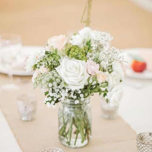 A bouquet of pale pink and white roses with baby's breath in a jar centerpiece, surrounded by votive candles on a table.