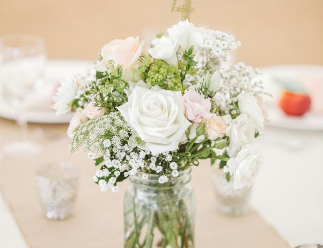 A bouquet of pale pink and white roses with baby's breath in a jar centerpiece, surrounded by votive candles on a table.