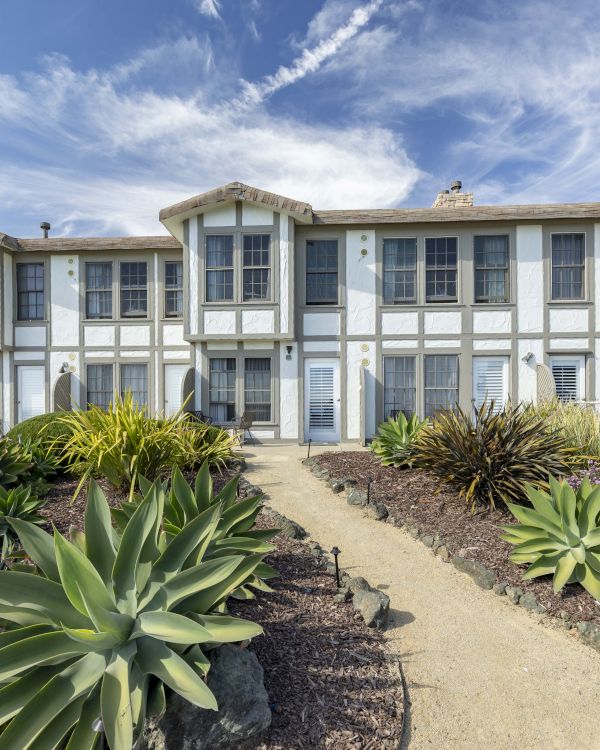 A white two-story apartment with arched dormers and windows, surrounded by landscaped succulents and purple flowers under a blue sky.