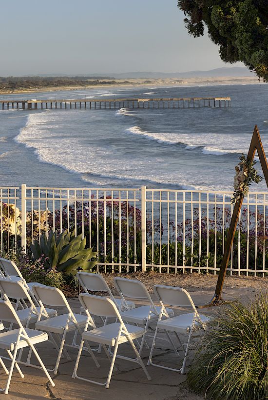 Oceanfront view with a long pier, waves rolling in, and empty lounge chairs on a terrace during sunset.
