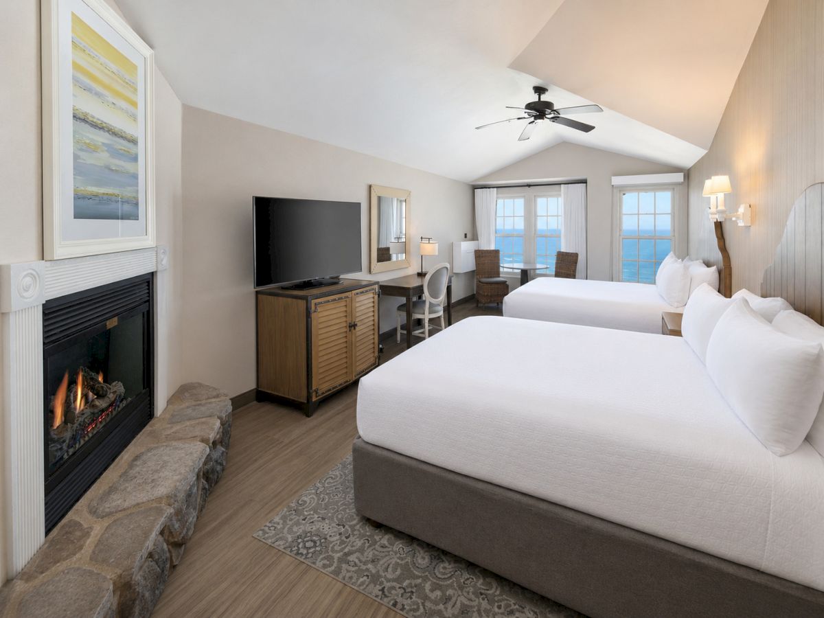 A bright hotel-style bedroom with two beds, a fireplace, flat-screen TV, and large windows showing the ocean.