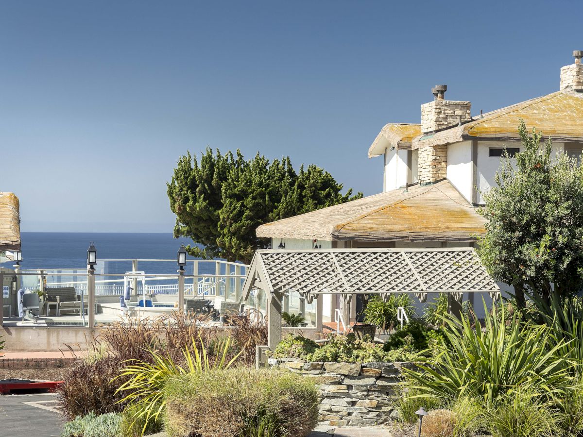 Rooftop terrace by the ocean with tiled roofs, stone chimneys, lounge furniture, and lush greenery under a clear sky.