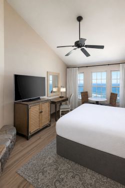 A bright hotel bedroom with a lit fireplace, TV on a console, large windows, and a ceiling fan above the bed.