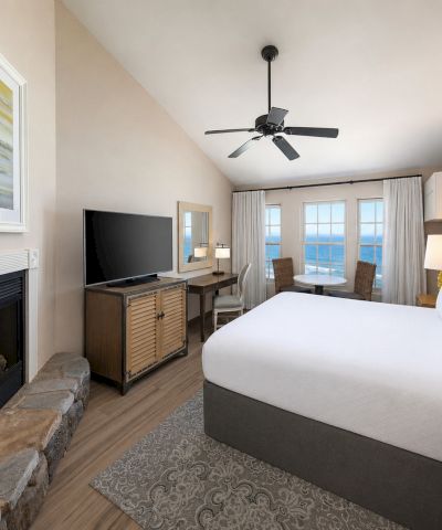 A bright hotel bedroom with a lit fireplace, TV on a console, large windows, and a ceiling fan above the bed.