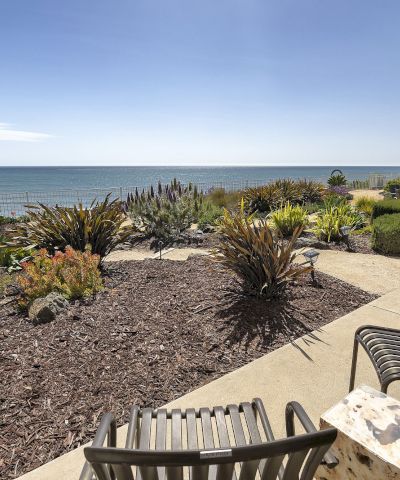 A sunny oceanfront patio with lush landscaped plants, a bench and table, and a wide view of the calm sea.