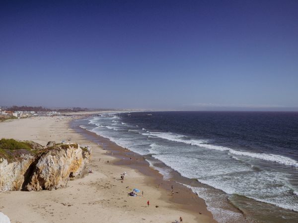 A wide sandy beach with people, gentle waves, rocky cliffs, and a long pier stretching along the coastline under a hazy sky.