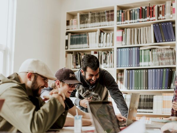In a library, a group of students huddle around a laptop, laughing and studying together among tall bookshelves.