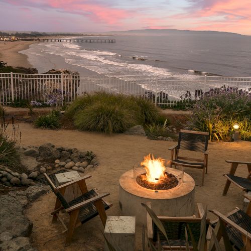 A beachfront sunset view with a pier, sandy shore, and patio chairs around a glowing fire pit by the ocean, ending the evening.