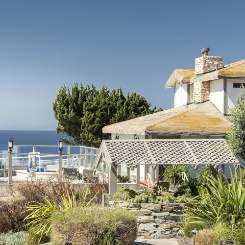 A sunny seaside terrace with white stone cottages, chimneys, lush shrubs, and a wide ocean view under a clear sky.