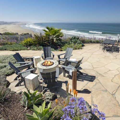 A seaside patio with lounge chairs around a roaring fire pit, facing a wide sandy beach and ocean beyond the railing.