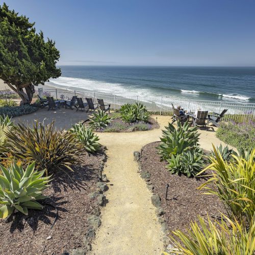 A seaside garden patio with chairs and lush plants overlooks waves and a sandy beach under a clear blue sky.