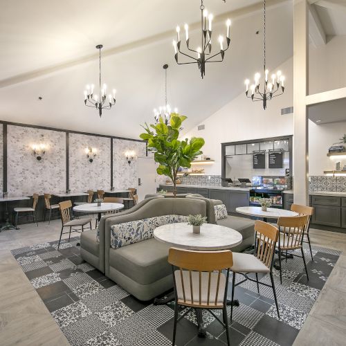 A modern caf&eacute;/lounge with velvet seating, round tables, and a bar counter under hanging chandeliers and bright decor.