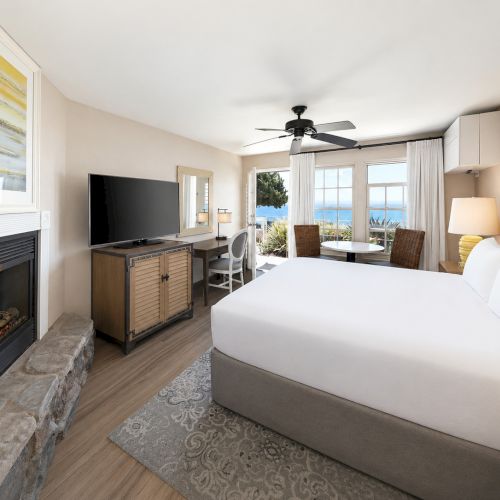 Bright hotel-style bedroom with a lit fireplace, TV, ceiling fan, and ocean-view windows.