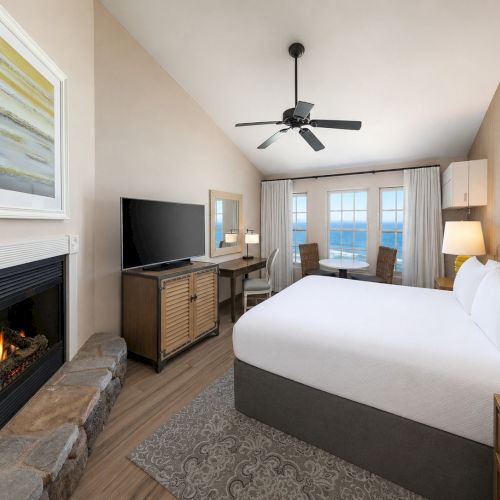 A cozy hotel bedroom with a king bed, wall TV, lit fireplace, and a ceiling fan above the seating area.