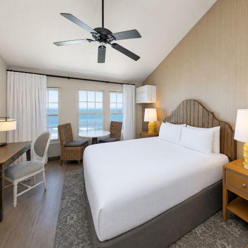 A bright hotel bedroom with a large bed, bedside lamps, a ceiling fan, and a window view of the ocean outside.