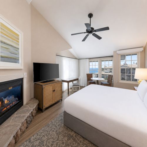 A bright hotel bedroom with a large bed, fireplace, flat-screen TV, ceiling fan, and ocean-view windows.