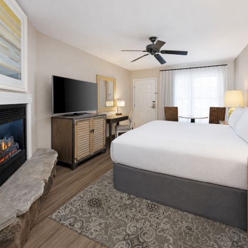 A modern hotel bedroom with a fireplace, flat-screen TV, ceiling fan, and made-up bed by a window.