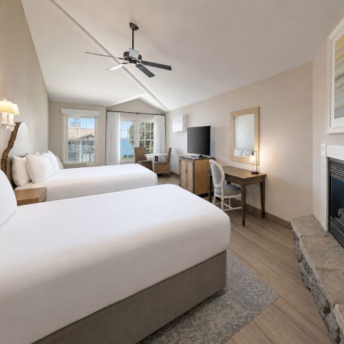 A bright hotel room with two beds, a ceiling fan, desk and TV, windows, and a lit fireplace.